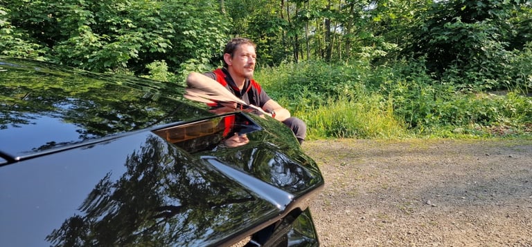 Man in red and black jacket installing a roof rack on a dark car against a wooded background